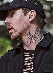 Close up of Professor Green looking to one side wearing blue and cream striped t-shirt, black collared jacket and black cap.