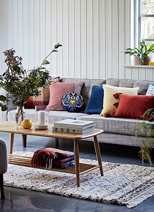 A grey sofa with a selection of different coloured cushions on top, behind a wooden coffee table topped with books, a folded throw, two candles and a vase full of flowers