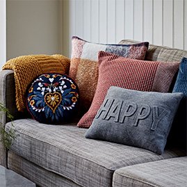 A grey sofa with a yellow knitted throw over one arm and a selection of blue, red and yellow cushions on top