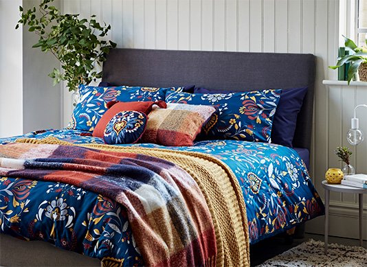 A double bed with a deep blue floral duvet set, a selection of blue, red and yellow cushions and two throws on top