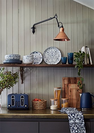 A kitchen counter and shelf with a selection of blue patterened crockery, a deep blue toaster and kettle set, clear food storage containers, wooden chopping boards and more