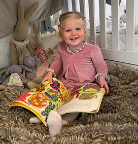 Small child wearing a pink dress in a fort made from her cot and a blanket, reading a book