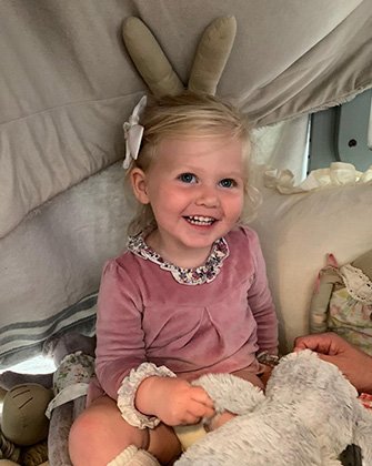 Small child wearing a pink dress in a fort made from her cot and a blanket, surrounded by teddies