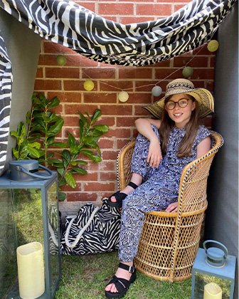 Child in a blue jumpsuit and sunhat sitting on a rattan chair in an outdoor blanket fort, complete with decorative string lights, candles and plants