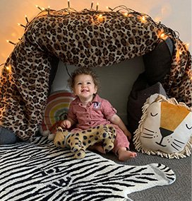 Small childing wearing a dusty pink jumpsuit sitting in a fort made from a cheetah print blanket, decorated with string lights, a lion cushion and a cheetah teddy