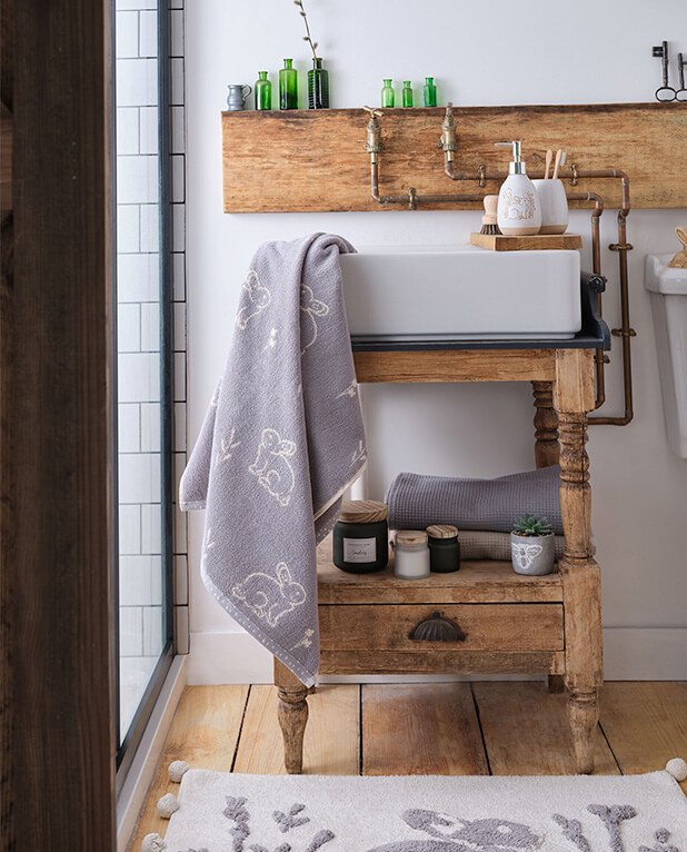 Rectangular white basin sink on wooden stand with bunny patterned towels, dispenser, tumbler, candles and artificial plant with wooden shelf in the background topped with green bottles and two large silver-tone keys.