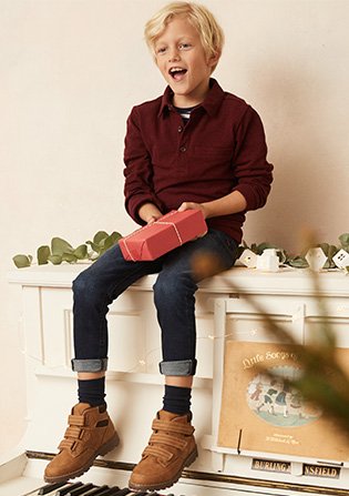A boy sitting on top of a piano holding a present wearing a burgundy jumper, dark wash jeans and brown boots.