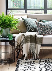 Room with large windows containing white sofa with Green Leaf Embroidered Cushions, Just Wellness Natural Yellow Contrast Large Piped Cushion, Green Checkered Throw, Cream Linear Berber Rug, black wire side table topped with artificial plant.
