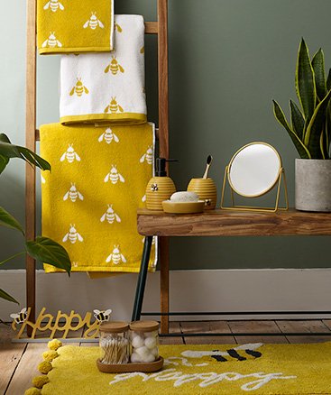 Wooden towel rack with yellow bee print towels, a wooden table topped with yellow bee tumblers and dishes, yellow round mirror and artificial plant, and a yellow bee happy bath mat.