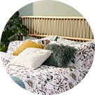 Double bed with wicker headboard with Floral Garden Easy Care Reversible Duvet Set, Green Mongolian Faux Fur Cushion, White Cushion and Yellow Cotton Velvet Large Cushion with large artificial plant to the side.