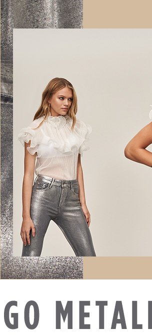 Woman with dark blonde hair poses wearing metallic silver trousers and white sheer short sleeve blouse with frill detailing.