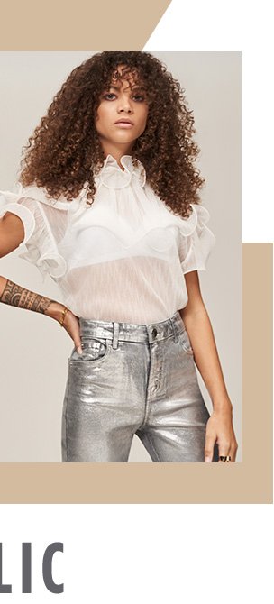 Woman with curly brown hair poses with hand on hip wearing metallic silver trousers and white sheer short sleeve blouse with frill detailing.