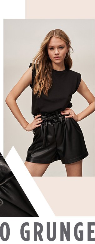Woman with dark blonde hair poses with hands on hips wearing black short sleeve top tucked into black PU paper bag belted shorts.