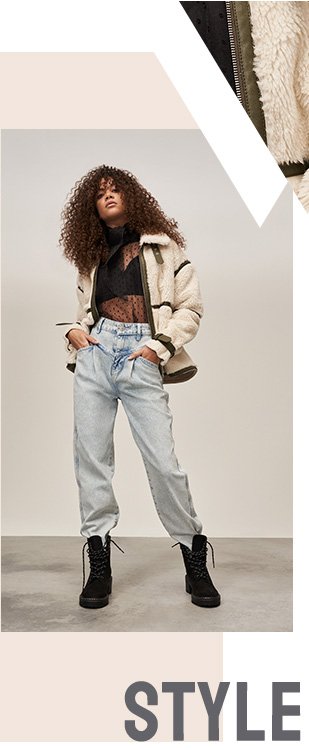 Woman with dark curly hair poses wearing black polka dot mesh collared top, cream and khaki sherpa jacket, acid wash jeans and black biker boots.