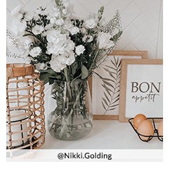 A glass vase full of white flowers next to a copper candle holder and an assortment of photo frames