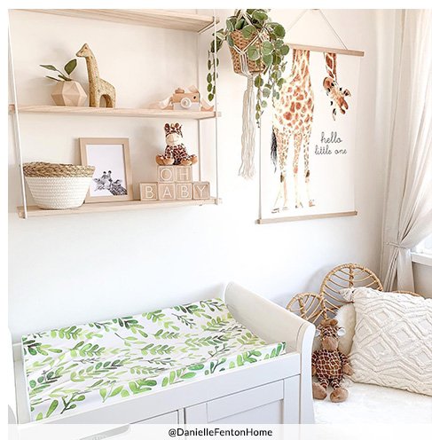 A baby's nursery with a white wooden changing station with a leaf print topper, decorated with a hanging plant, white ruffle cushions and a shelf with a selection of giraffe and tiger toys