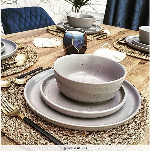 A dining table set with grey crockery, black and gold cutlery, woven placemats and a blue accent candle holder