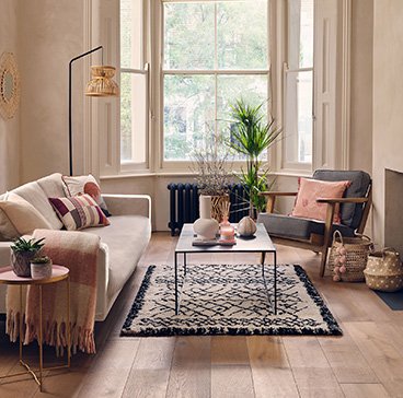 Living room with large open windows featuring cream sofa, grey chair with pink and cream scatter cushions, coffee table, cream textured rug and rattan floor lamp.