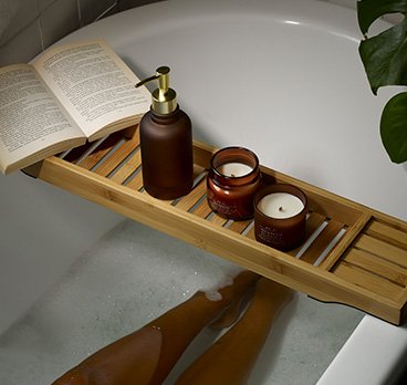 Legs of a woman in a bath with a wooden bath tray topped with an amber soap dispenser, 2 amber candles and an open book.
