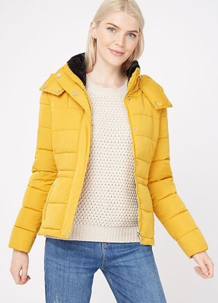 A woman wearing a yellow high collar padded jacket over a cream knit jumper and blue jeans. 