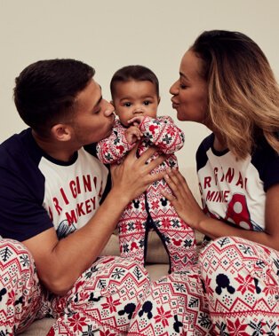 Man and woman wearing Disney Mickey Mouse matching family Christmas pyjamas lean in to kiss baby wearing matching sleepsuit.