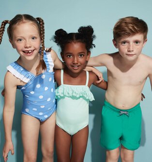 Three children standing shoulder to shoulder wearing a variety of swimming costumes and trunks