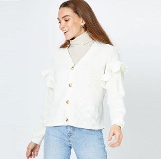 A smiling woman wearing an ivory ribbed frill sleeve cardigan over a neutral roll neck top and blue jeans.