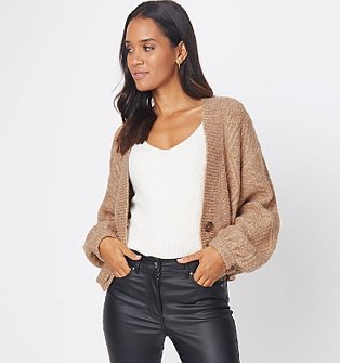 A woman wearing a tan cable knit cardigan over a white vest and black trousers.