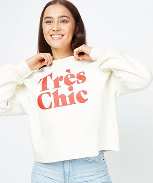 Woman poses smiling wearing cream très chic slogan sweatshirt and light wash blue jeans.