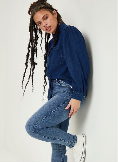 A woman posing in mid wash denim jeans, white trainers and a dark wash denim shirt.