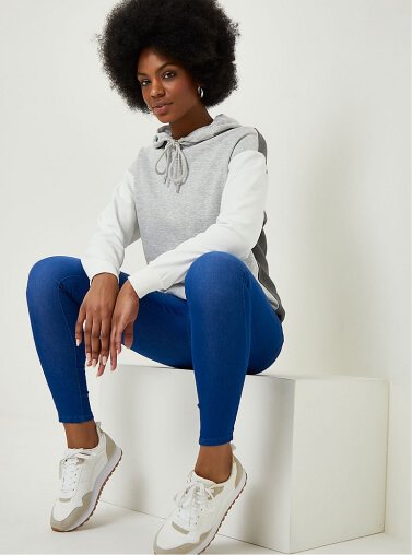 A woman sitting down in jeggings, white trainers and a grey panel hoodie.