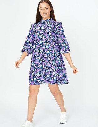 Woman poses wearing purple floral print high neck dress and white trainers.