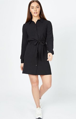 Woman poses wearing black long sleeve mini shirt dress and white trainers.