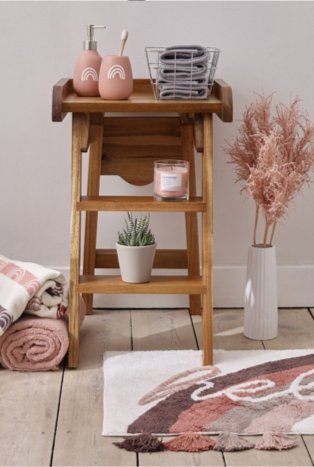 Wooden-effect bathroom cabinet features pink rainbow-patterned tumbler and dispenser set, wire basket containing grey face cloths, pink candle and artificial plant with cream rainbow-patterned towel, pink towel and cream hello slogan tassel trim bathmat on the floor.