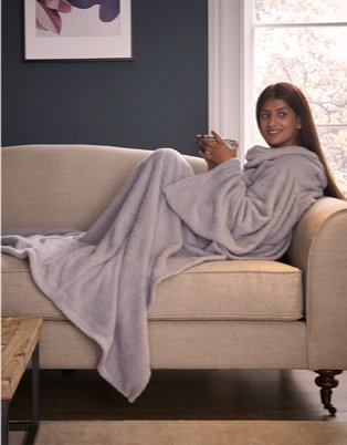 Woman sits on cream sofa under lilac blanket holding mug.