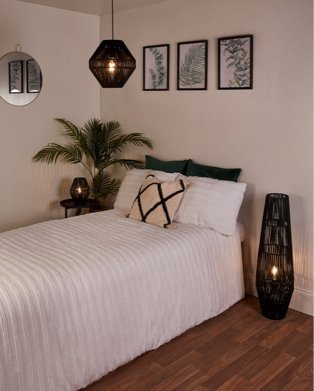 Room features double bed with white bedding, green pillows and natural diamond-patterned textured cushion, round wall mirror, 3 framed leaf prints, black rattan floor lamp, black rattan lampshade and artificial plants.