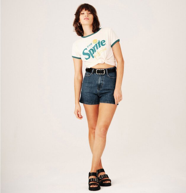 A woman posing in a Sprite slogan t-shirt, denim shorts and black sandals.