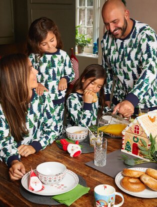Family wearing matching tree print Christmas pyjamas building a gingerbread house.
