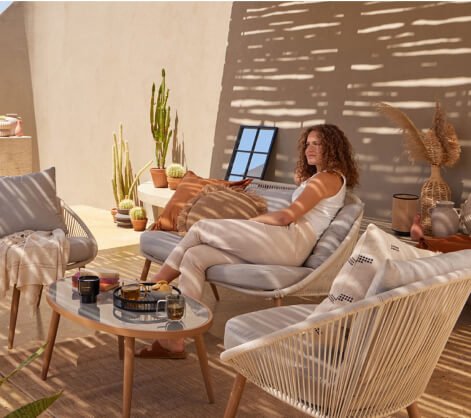 Woman sits on wicker sofa set surrounded by assorted garden accessories.