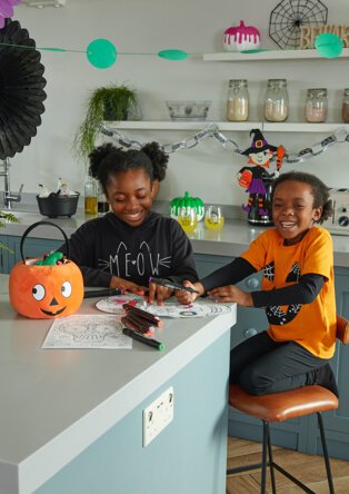 Little girl and boy celebrating Halloween at home by colouring in Skeleton mask, girl wearing a black t-shirt with the words ‘Meow’ and boy wearing an orange t-shirt with black sleeves with a pumpkin face on