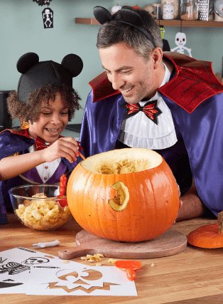 A parent and child carving pumpkins in Mickey Mouse Vampire costumes.