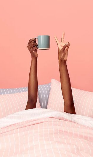 Pink duvet set with woman’s hands pointing upwards holding a grey mug in right hand and gesturing the peace sign with the left.