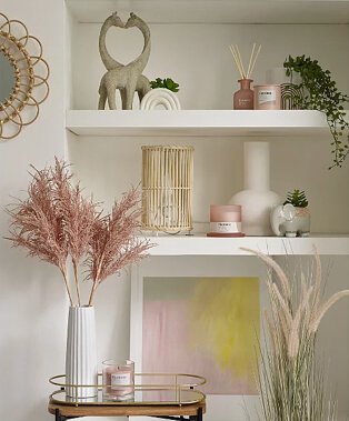 Two shelves topped with giraffe ornament, reed diffuser, rainbow artificial plant, wicker lamp, white vase, elephant artificial plant and green, pink and yellow framed print.