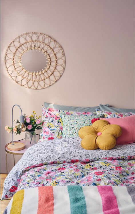 A colourful floral duvet set, striped blanket and assortment of bright cushions on a double bed, a gold bedside table topped with flowers and a lamp, and a wicker mirror on the wall.