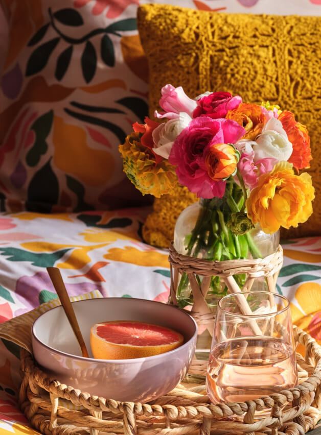 A glass vase with orange, yellow and pink flowers, grapefruit in a pale pink bowl and a glass of water on a wicker tray on a bed.