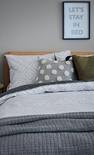 Double bed with wooden headboard and white and grey printed duvet, grey textures throw and grey and white polka dot and olive-green scatter cushion with black framed 'let's stay in bed' print hanging above.