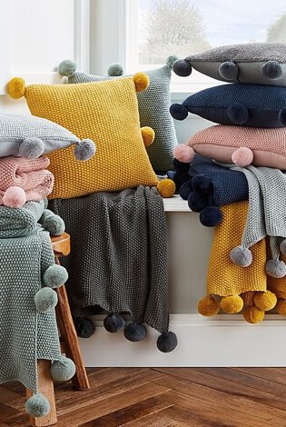 Bright room with large window, white walls and wooden floor with layers of grey, pink, mustard and navy pom-pom cushions and throws.