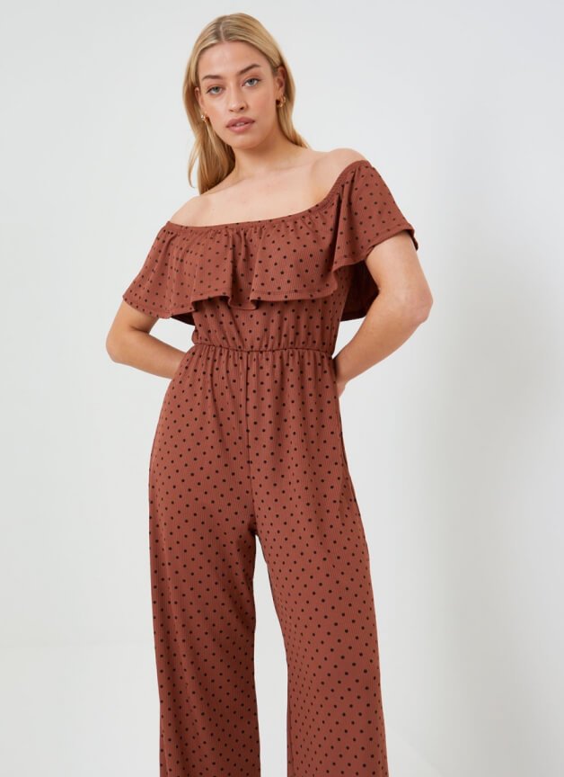 A woman posing in a brown polka dot off the shoulder jumpsuit.