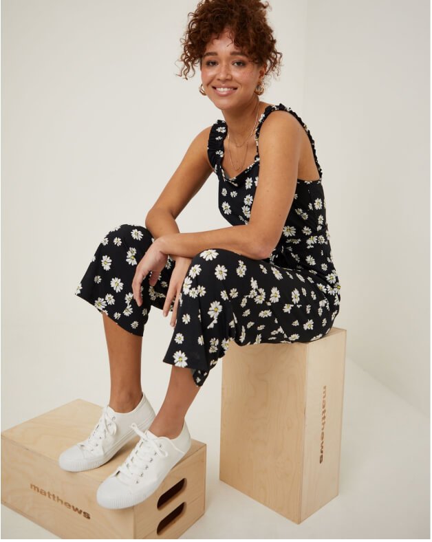 A woman sitting on a box wearing a black daisy print jumpsuit and white trainers.