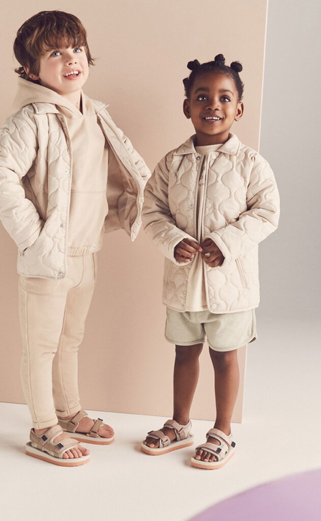 Two children wearing matching cream outfits, including quilted jackets, hoodies, shorts, trousers and sandals.